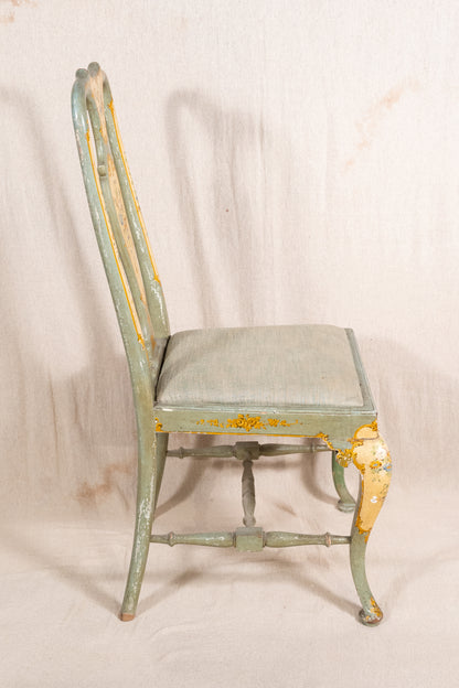 A distressed vintage wooden dining chair with a light sage green painted finish and peeling yellow-gold paint, featuring a padded seat cushion, photographed against a neutral off-white fabric backdrop.