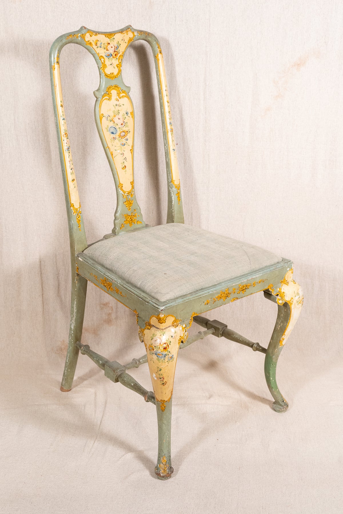 An antique-style wooden dining chair with a distressed sage green and yellow painted finish, featuring a high curved backrest and a cushioned seat, set against a neutral fabric backdrop.