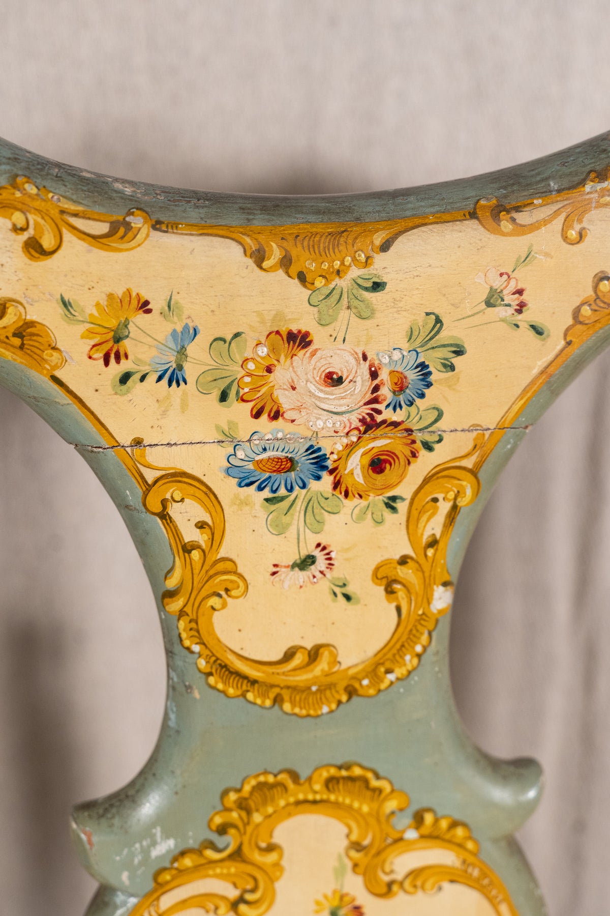 Close-up of an ornate vintage chair back featuring a pale sage green painted wood frame with intricate gold gilding and a central floral motif in yellow, blue, red, and green.