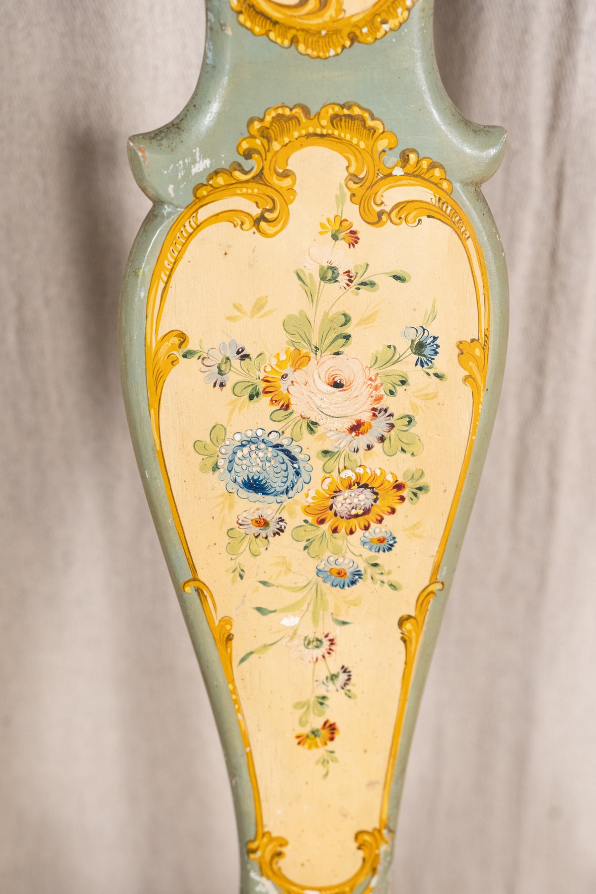 Close-up of an ornate vintage-style chair back or panel, featuring a pale sage green painted frame, a cream center panel, and hand-painted floral motifs in blue, yellow, and orange with gold accents.