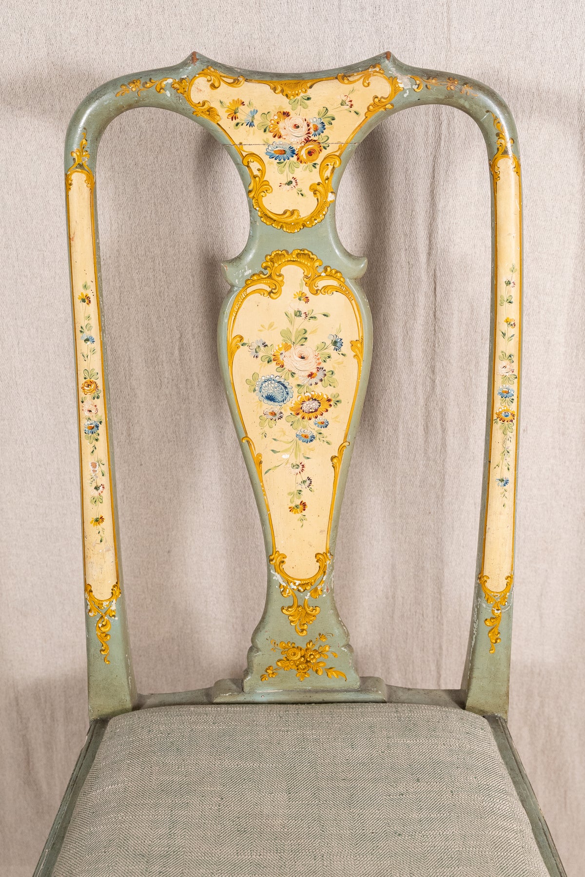 An ornate vintage-style chair back with a pale sage green painted frame, cream-colored panels, and intricate gold floral and scrollwork detailing, set against a neutral draped fabric backdrop.