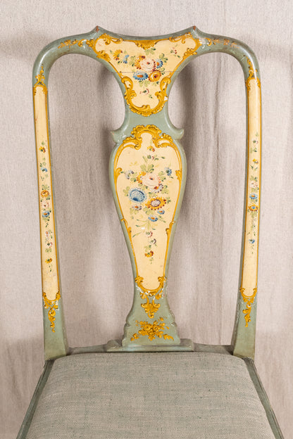 An ornate vintage-style chair back with a pale sage green painted frame, cream-colored panels, and intricate gold floral and scrollwork detailing, set against a neutral draped fabric backdrop.