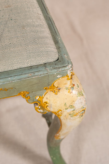 Close-up of an antique upholstered chair corner showing a distressed sage green painted frame, gold gilded cabriole-style legs, and a gray herringbone fabric seat cushion.