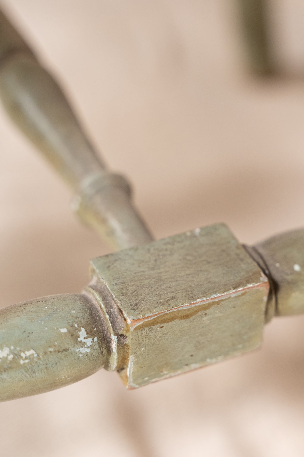 Close-up of a distressed, weathered metal or wood furniture joint with a muted sage green and brown patina, set against a soft beige background.