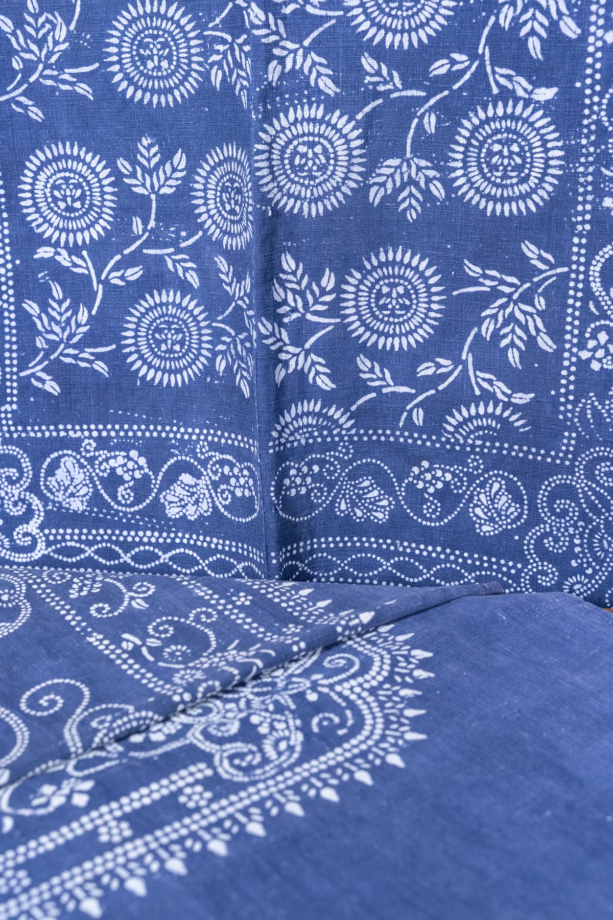 A close-up of a blue fabric with a white hand-block printed floral and paisley pattern, showing the texture and detailed stitching.
