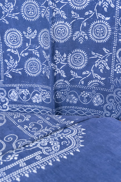 A close-up of a blue fabric with a white hand-block printed floral and paisley pattern, showing the texture and detailed stitching.