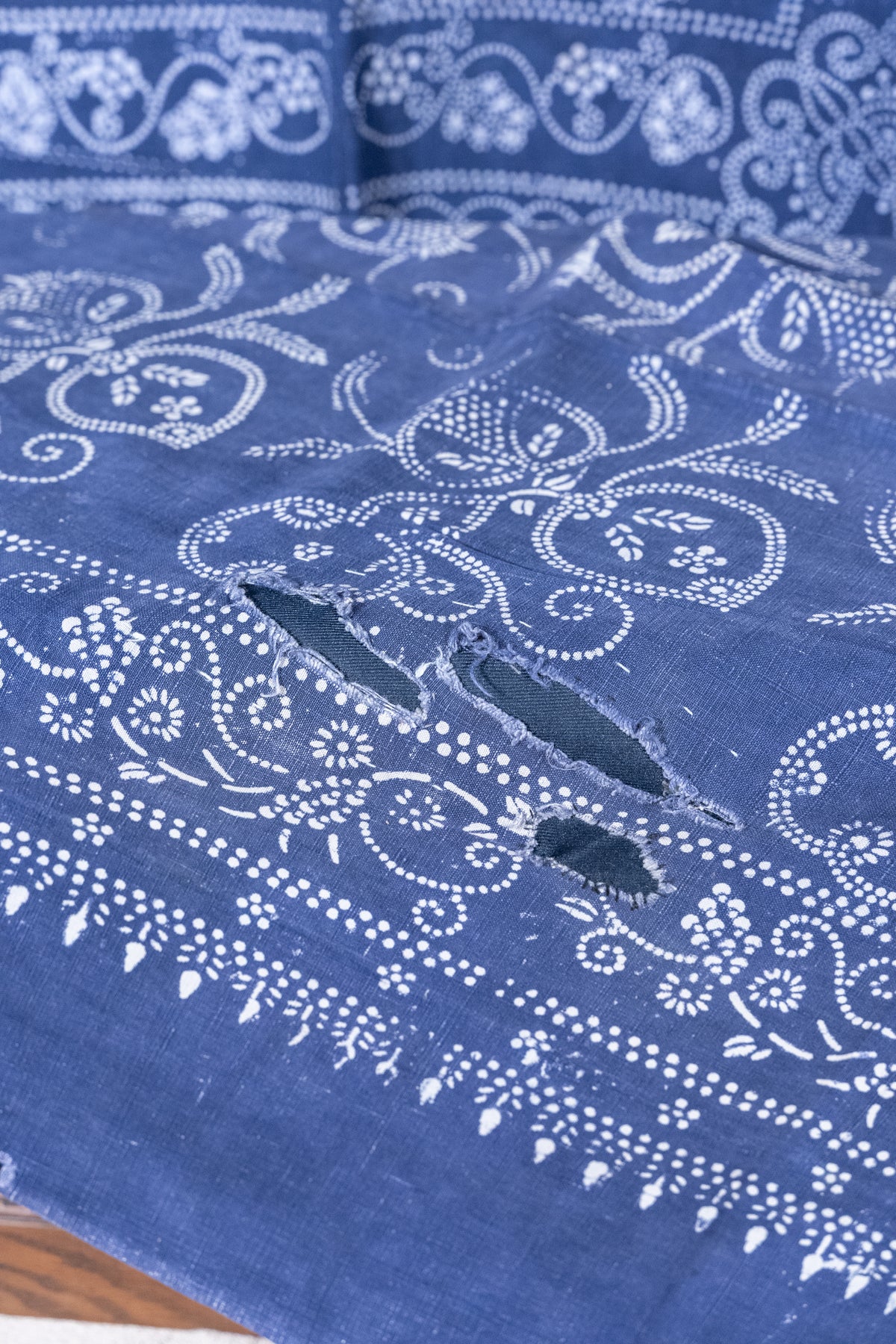 A close-up of a blue fabric with white floral and paisley bandhani-style prints and two distressed, raw-edge holes near the center.