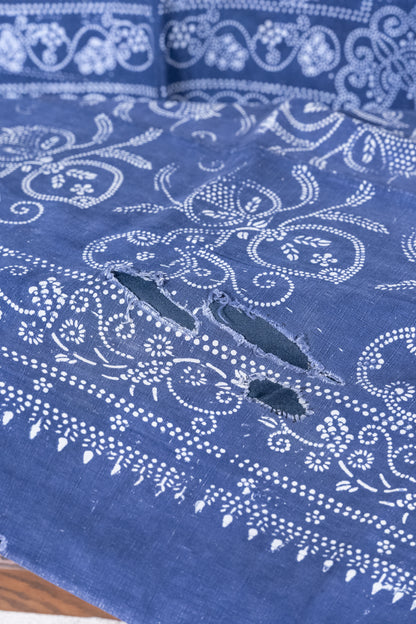 A close-up of a blue fabric with white floral and paisley bandhani-style prints and two distressed, raw-edge holes near the center.