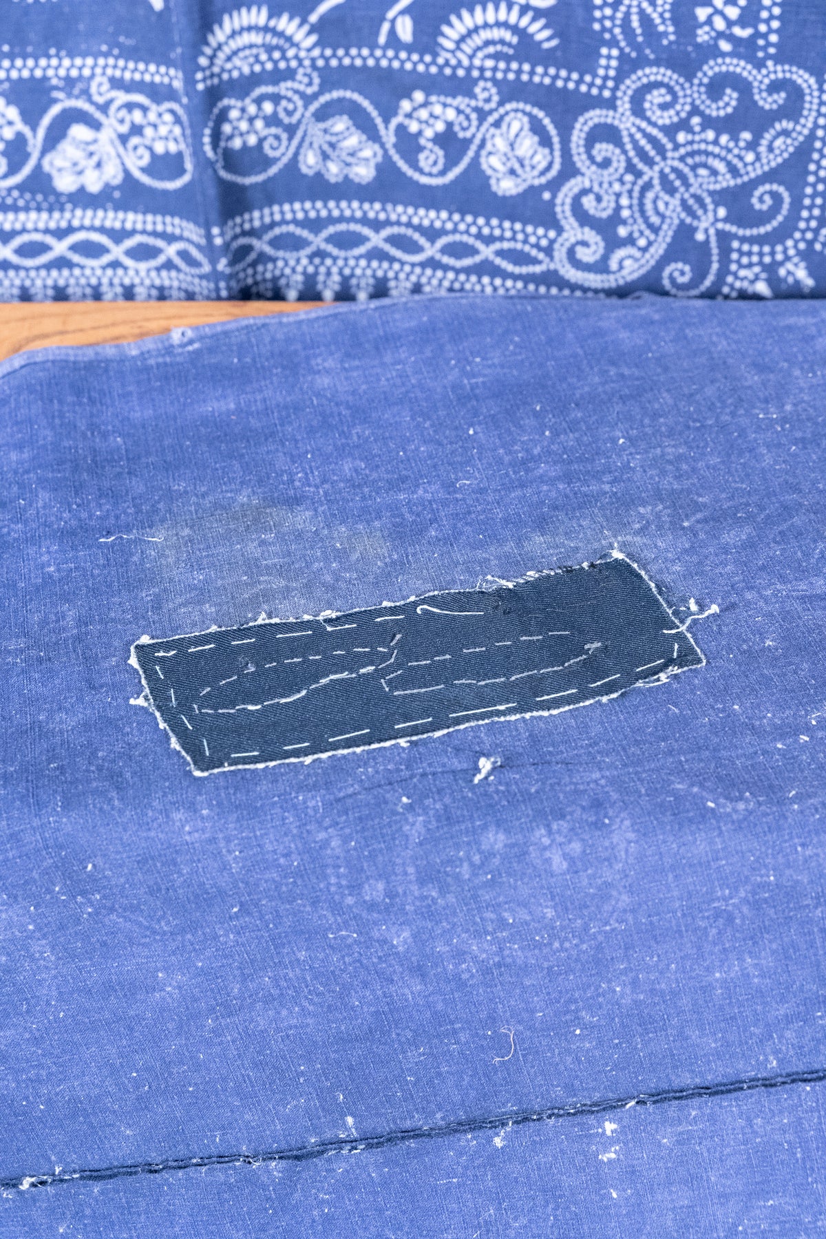 A close-up of a blue fabric surface with a rectangular patch of darker blue material sewn onto it, with a patterned blue and white textile visible in the background.