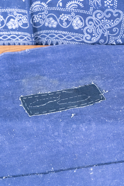 A close-up of a blue fabric surface with a rectangular patch of darker blue material sewn onto it, with a patterned blue and white textile visible in the background.
