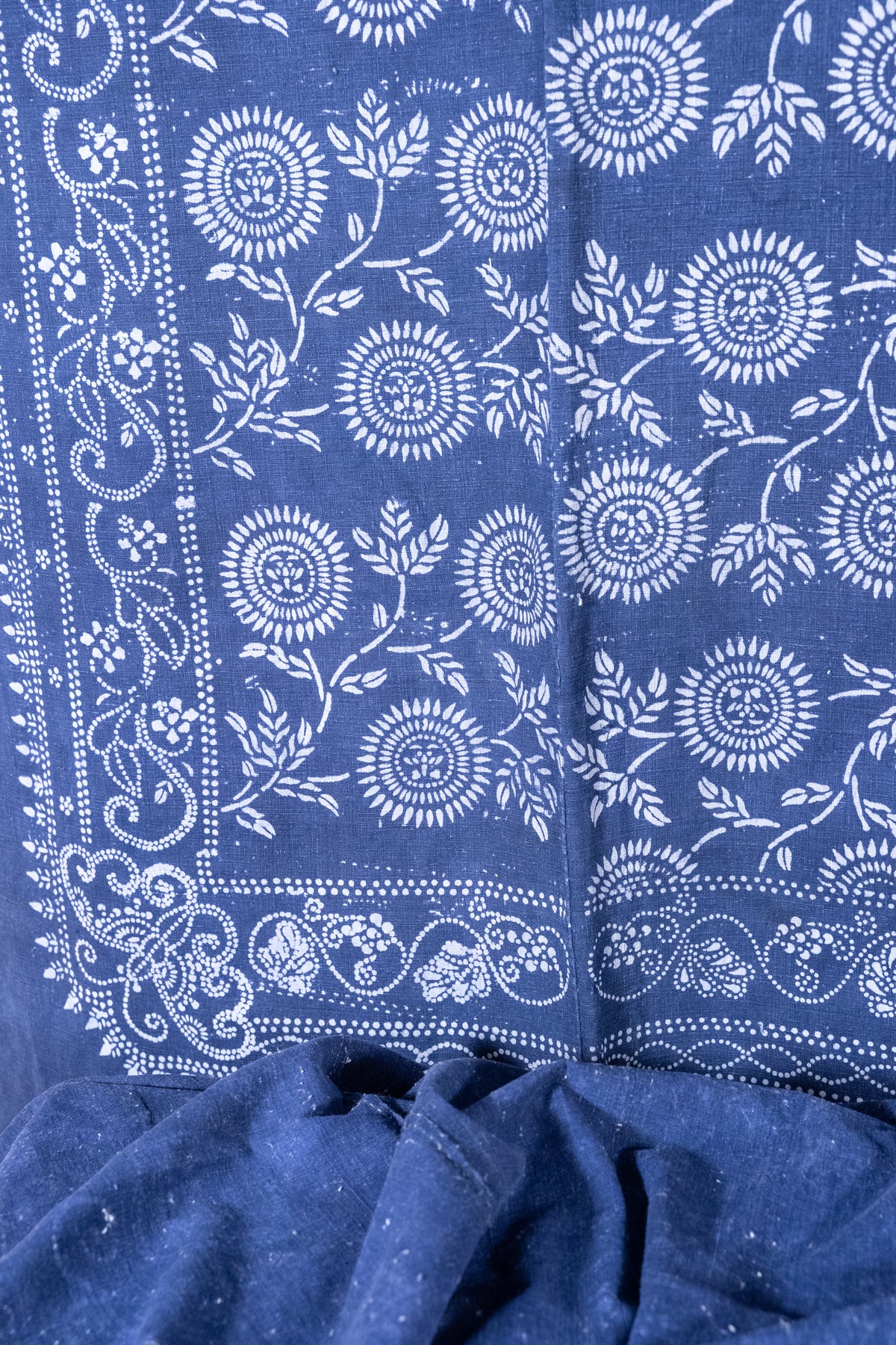A close-up of indigo blue fabric with white floral and vine block-print patterns, showing the textile texture and border design.