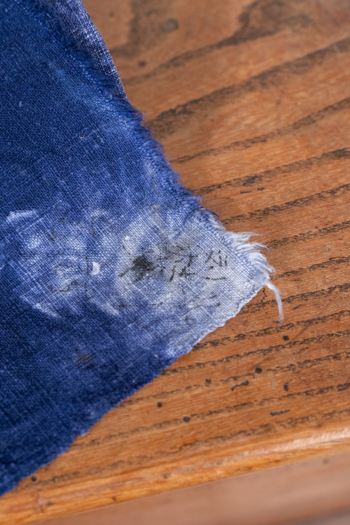 A close-up of a dark blue denim fabric swatch with a frayed, distressed edge, laid diagonally on a brown wooden surface.
