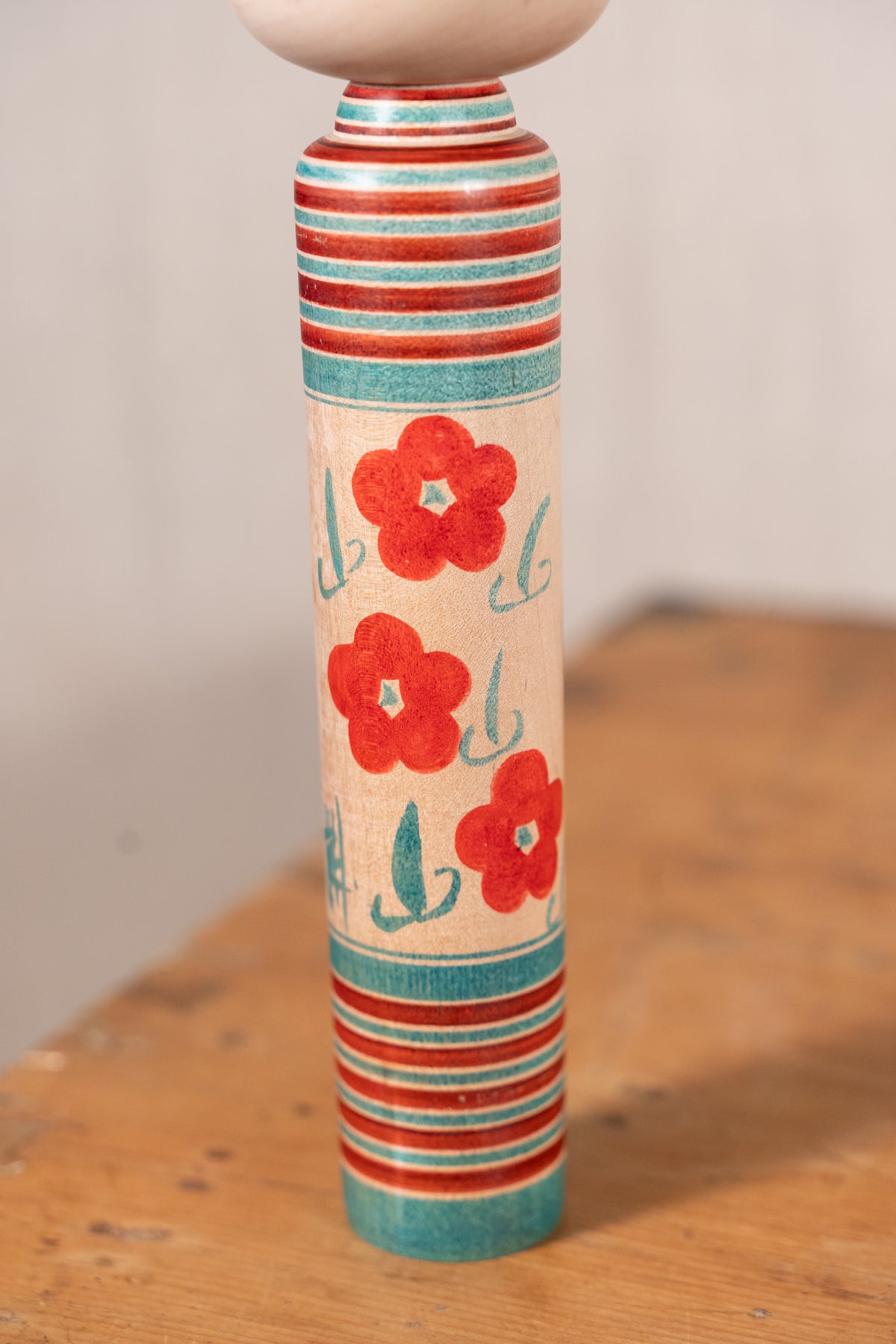 A hand-painted wooden spindle top featuring red flowers and teal stripes on a light wood background, standing on a rustic wooden table.