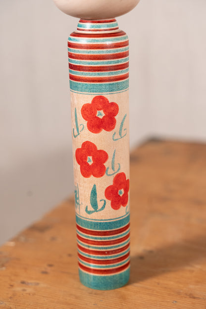 A hand-painted wooden spindle top featuring red flowers and teal stripes on a light wood background, standing on a rustic wooden table.