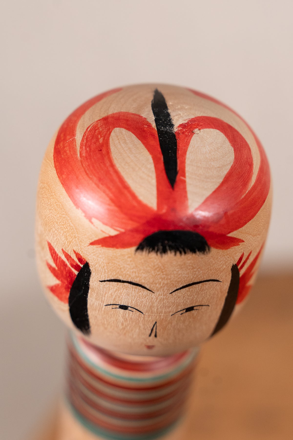 A close-up of a hand-painted wooden geisha doll head with red hair, black hair details, and a striped neck, set against a soft beige background.