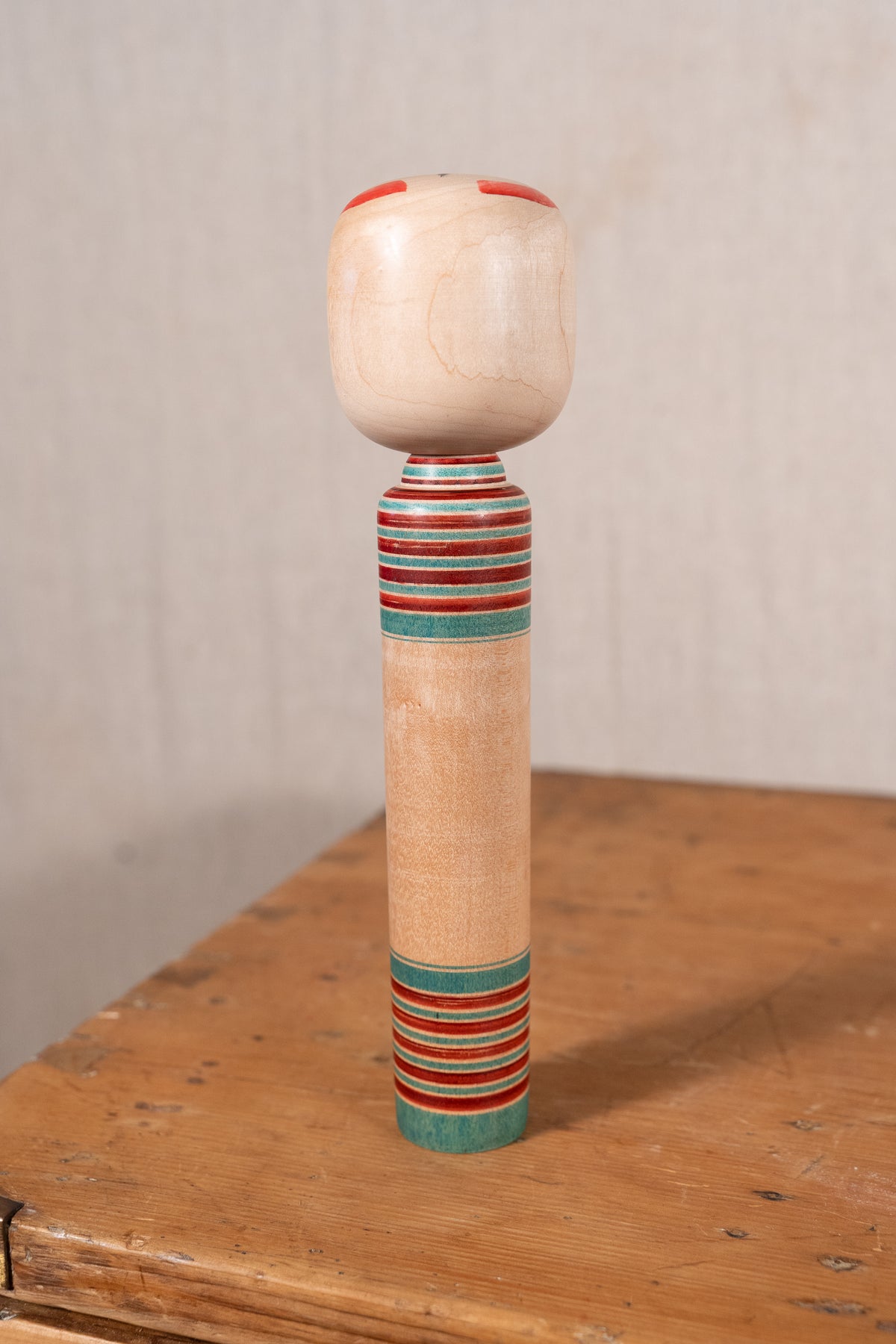 A handcrafted wooden spindle with a rounded natural wood top, decorated with red and teal painted stripes, standing on a rustic wooden table against a neutral beige background.