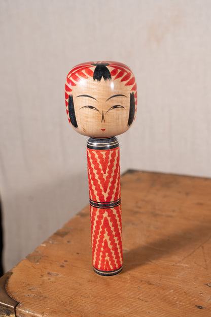 A traditional Japanese wooden doll or kokeshu standing upright on a rustic wooden table against a neutral beige wall.