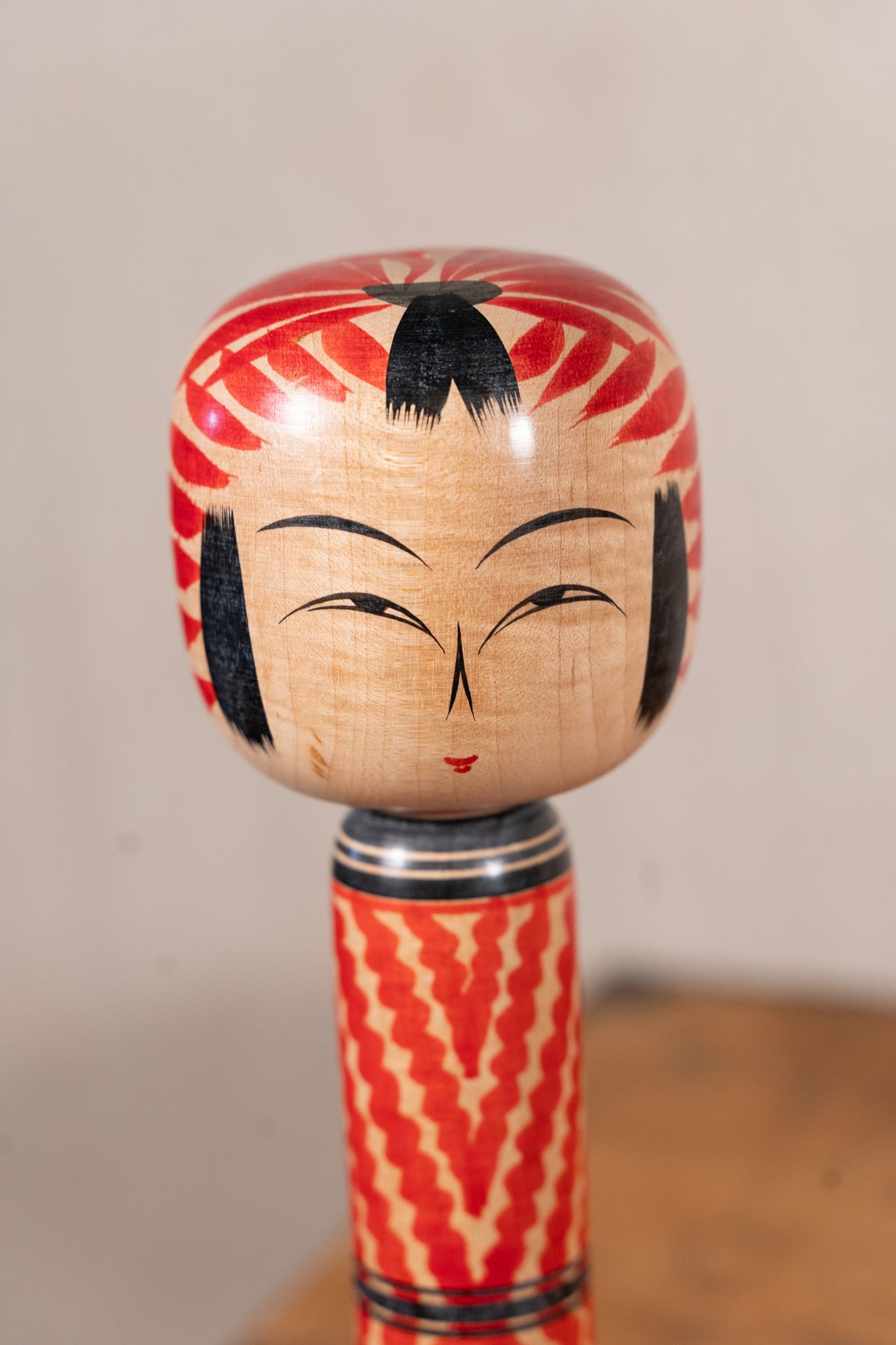 A traditional Japanese wooden doll (Kekakeboko) with a hand-painted face, red patterned hair, and a patterned wooden body, photographed against a neutral beige background.