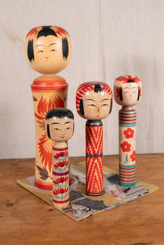Four hand-painted Japanese kokeshu dolls in different sizes standing on a small printed mat on a wooden table against a neutral background.