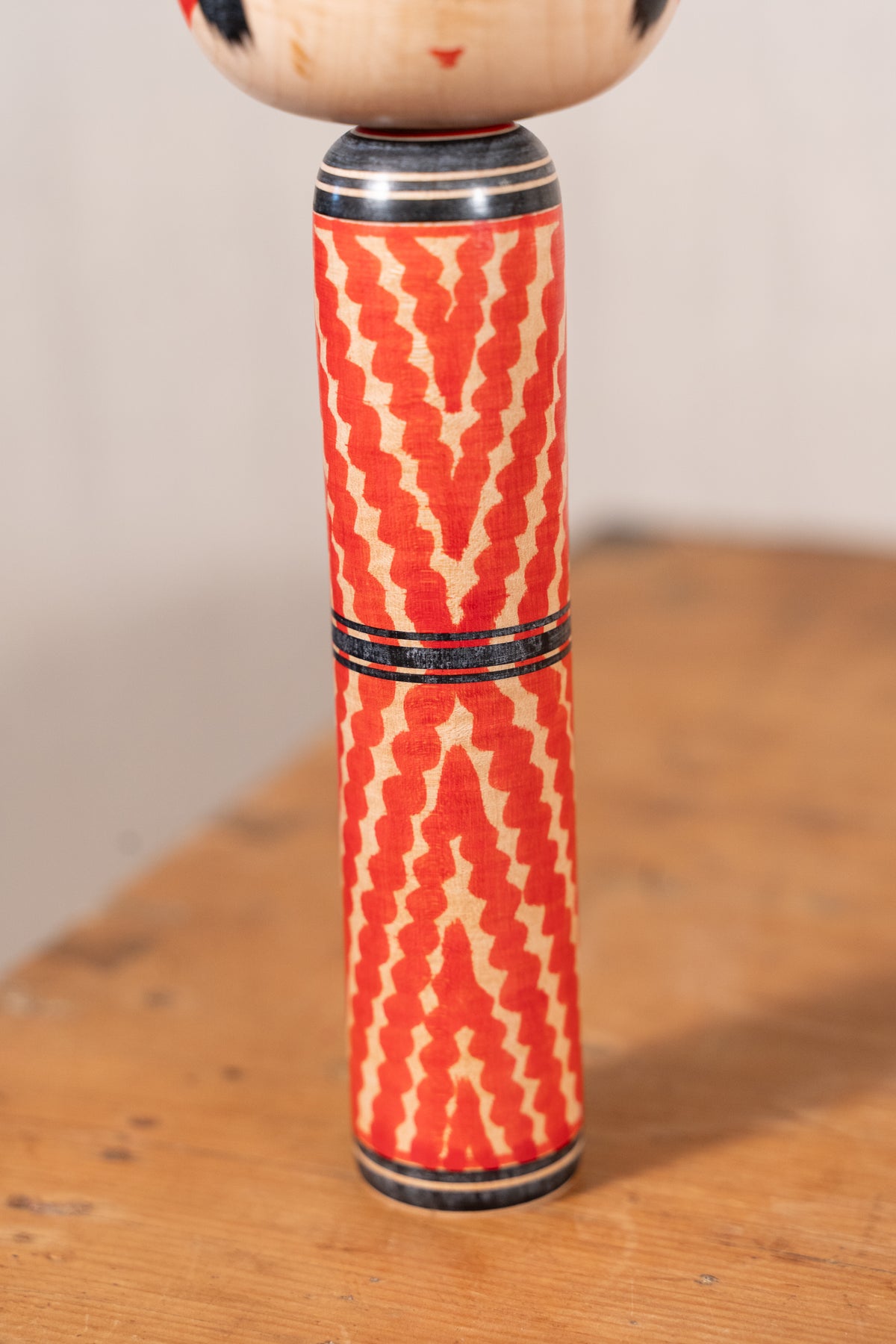 A close-up of a traditional Japanese lacquerware doll's cylindrical torso, featuring a red and beige geometric pattern with black painted bands, standing on a wooden surface.