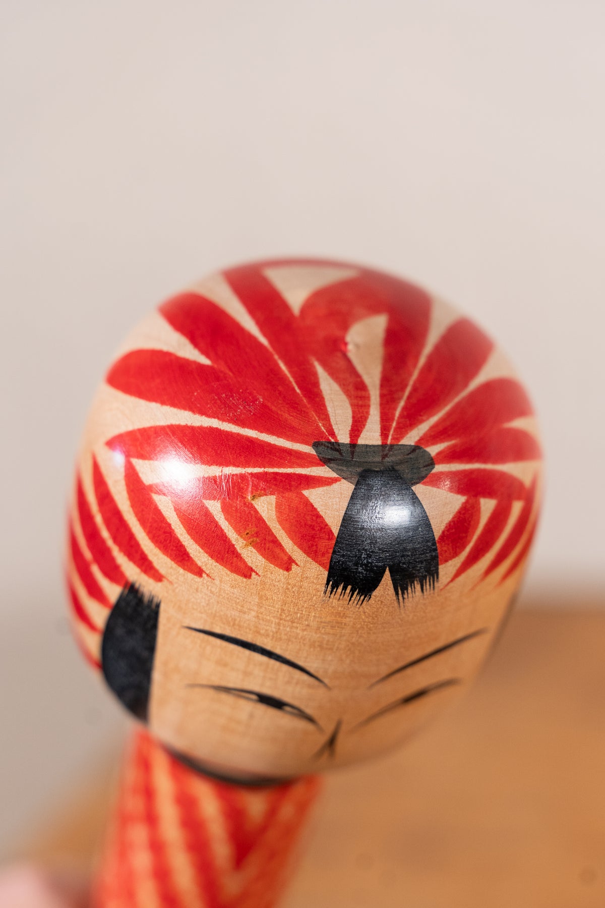 A close-up of a Japanese kokeshu doll head with a glossy red and beige striped hair pattern, black hair bow and face details, and a striped red and beige body, set against a soft beige background.