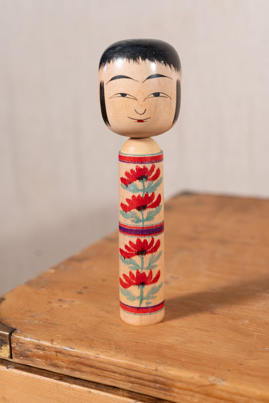 A traditional Japanese wooden doll (kayoi-kakemono) with a painted face and black hair, standing on a rustic wooden surface against a neutral beige background.