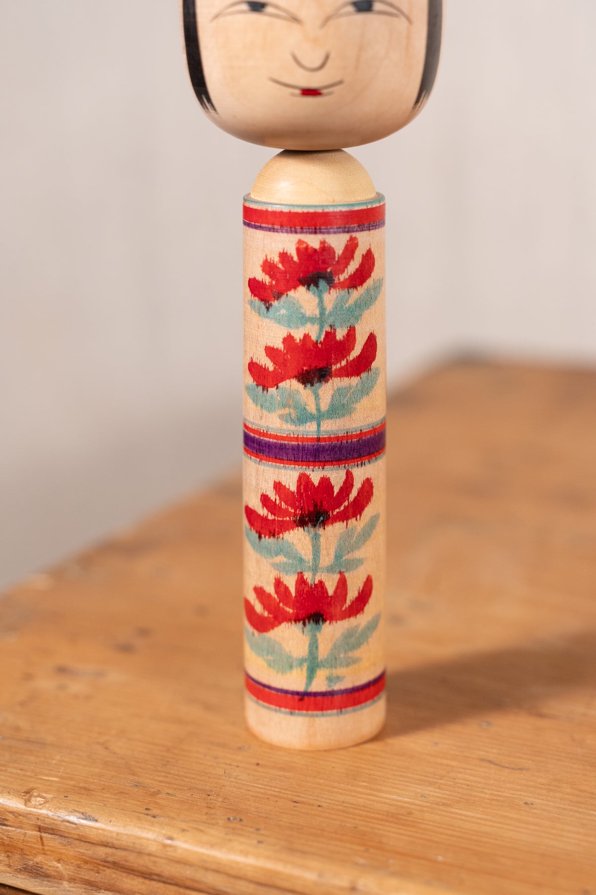 A handcrafted wooden kokeshu doll with a painted face and a decorated cylindrical body featuring red flowers and blue-green leaves, standing on a wooden surface.