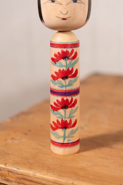 A handcrafted wooden kokeshu doll with a painted face and a decorated cylindrical body featuring red flowers and blue-green leaves, standing on a wooden surface.