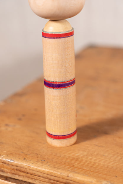 A close-up of a traditional wooden kokesh doll, showing its cylindrical body with red and blue woven stripes and a round wooden top, standing on a rustic wooden surface.