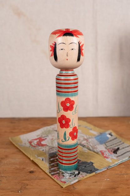 A hand-painted Japanese wooden doll (gekisori) with a decorated head and striped, floral-patterned body, standing on a vintage paper card over a wooden surface.