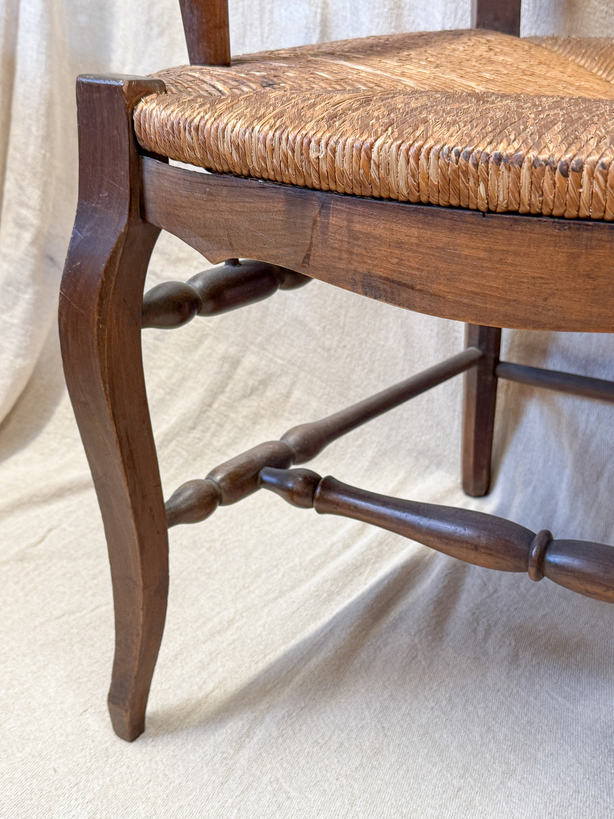 Close-up of a wooden dining chair with a woven rush or cane seat, showing the dark brown wood frame and turned spindles against a draped cream fabric backdrop.