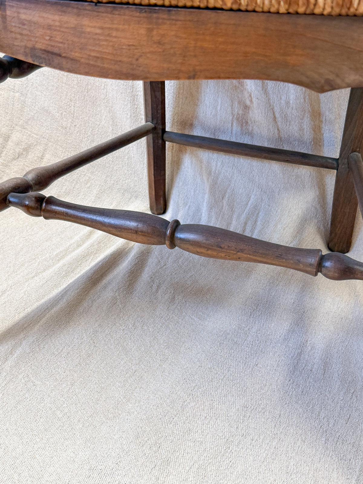 Close-up of a dark brown wooden chair leg with turned spindles and a woven seat cushion, photographed against a draped beige fabric backdrop.