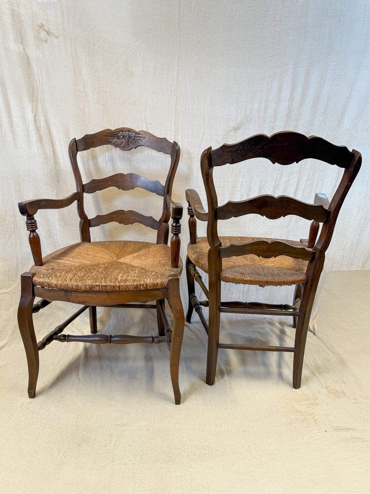Two dark wood ladder-back dining chairs with woven cane seats, shown side by side against a wrinkled off-white fabric backdrop.
