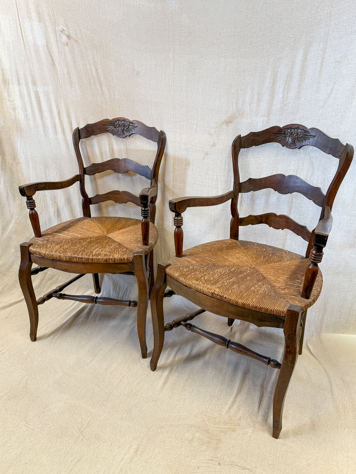Two vintage wooden ladder-back dining chairs with woven rush seats, shown against a draped off-white fabric backdrop.