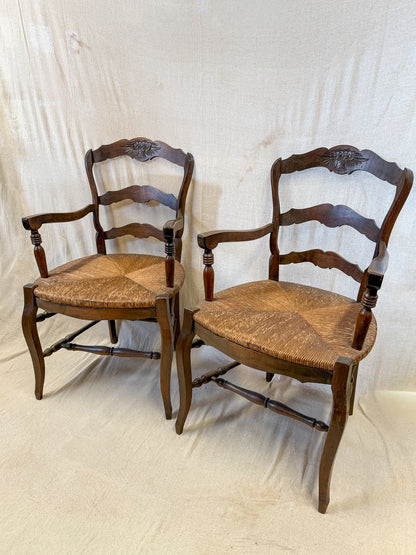 Two vintage wooden ladder-back dining chairs with woven rush seats, shown against a draped off-white fabric backdrop.