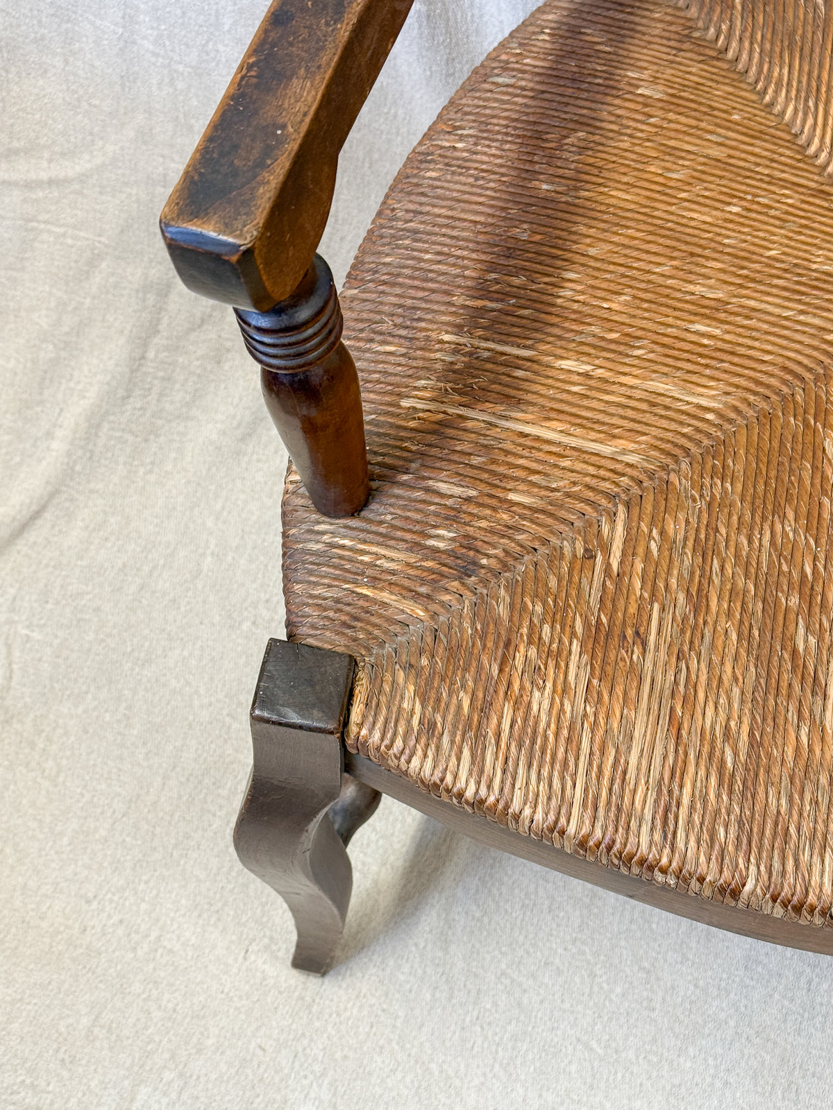 Close-up of a wooden chair showing the woven seat, turned wooden leg, and decorative metal corner brace on a light fabric background.