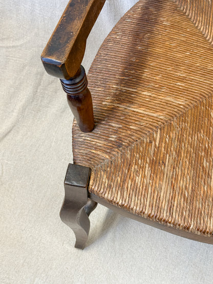 Close-up of a wooden chair showing the woven seat, turned wooden leg, and decorative metal corner brace on a light fabric background.