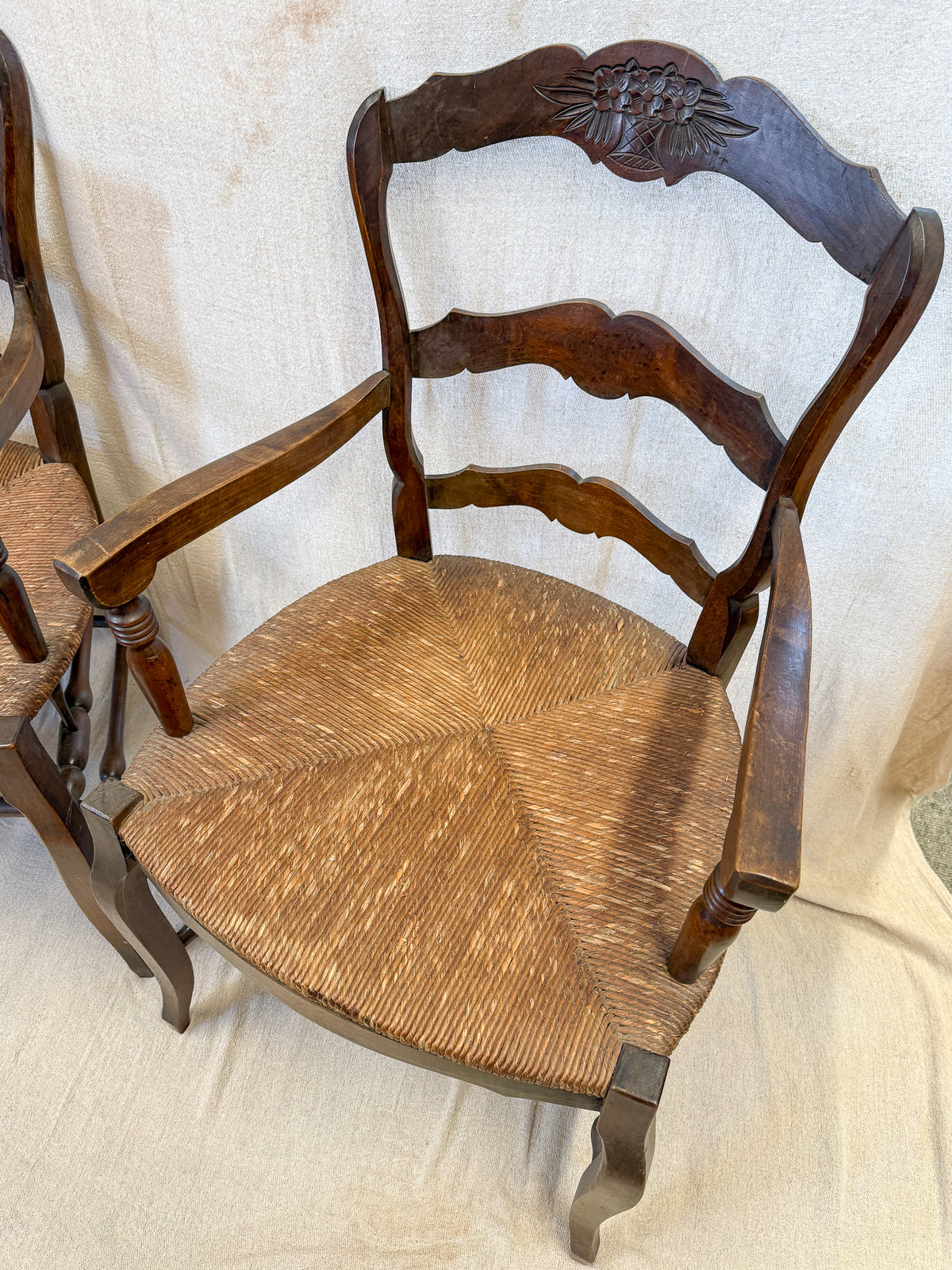 A dark brown wooden dining chair with a woven cane seat and a carved floral backrest, shown against a draped beige fabric backdrop.