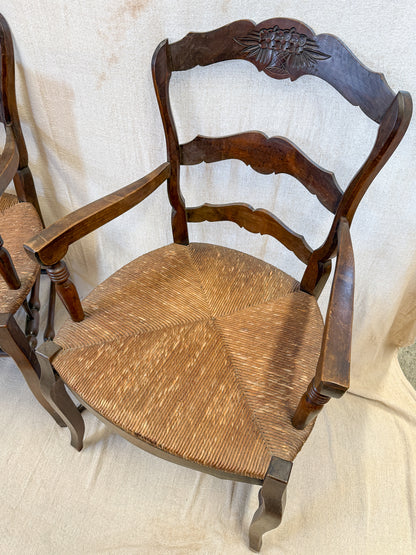 A dark brown wooden dining chair with a woven cane seat and a carved floral backrest, shown against a draped beige fabric backdrop.