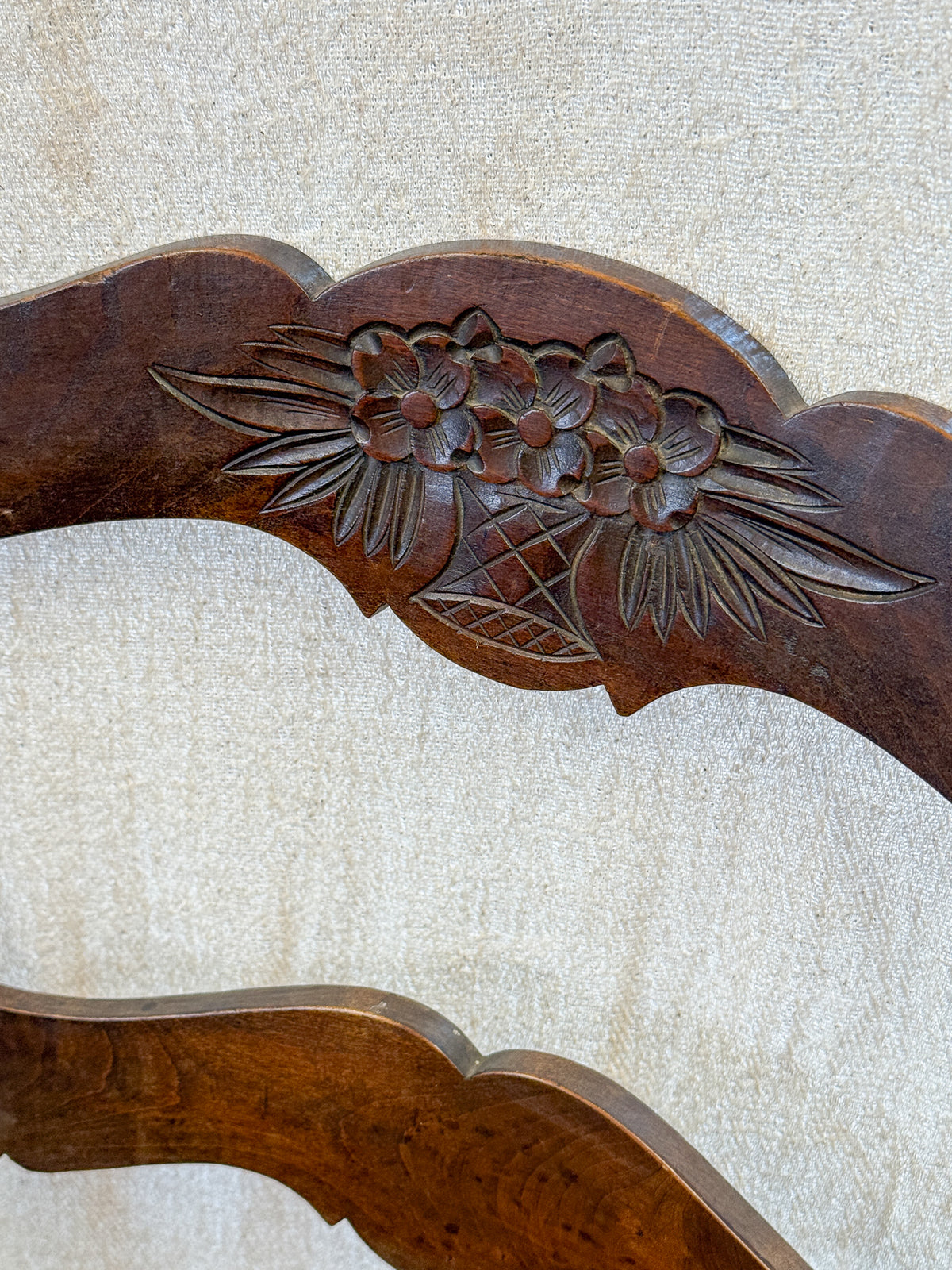 Close-up of a dark brown carved wooden chair back featuring a floral motif, set against a textured beige fabric background.