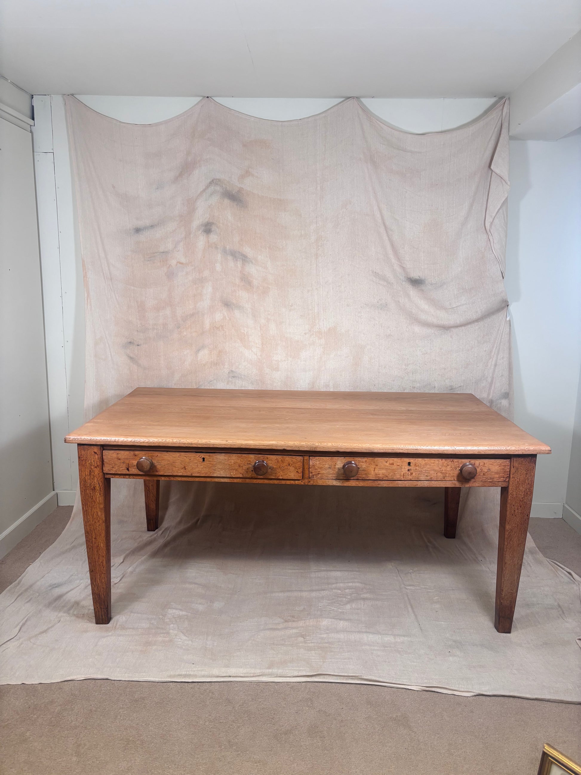 A rustic wooden dining table with three drawers and round metal knobs, set against a draped beige fabric backdrop in a room.