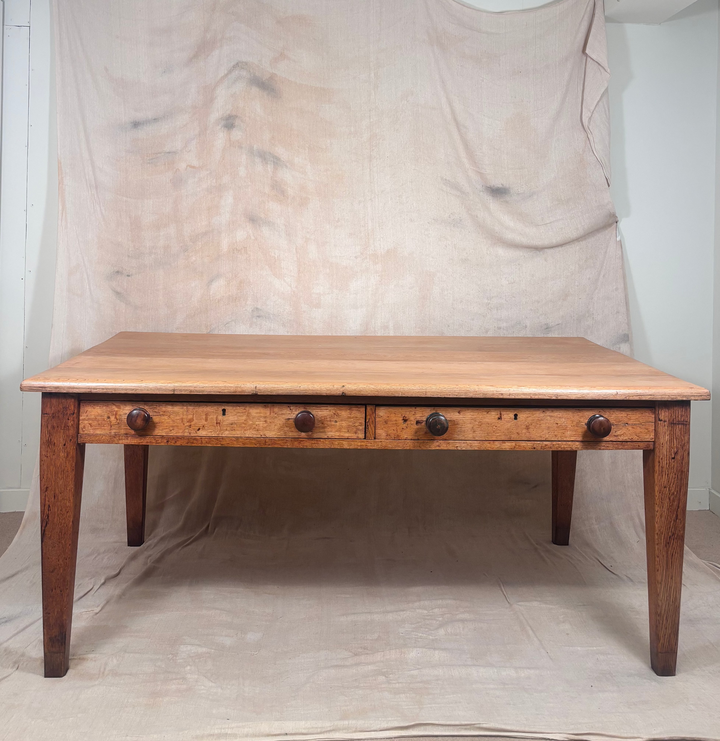 A rustic wooden dining table with three drawers and dark metal knobs, set against a backdrop of draped beige fabric.