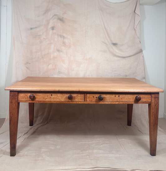 A rustic wooden dining table with three drawers and dark metal knobs, set against a backdrop of draped beige fabric.