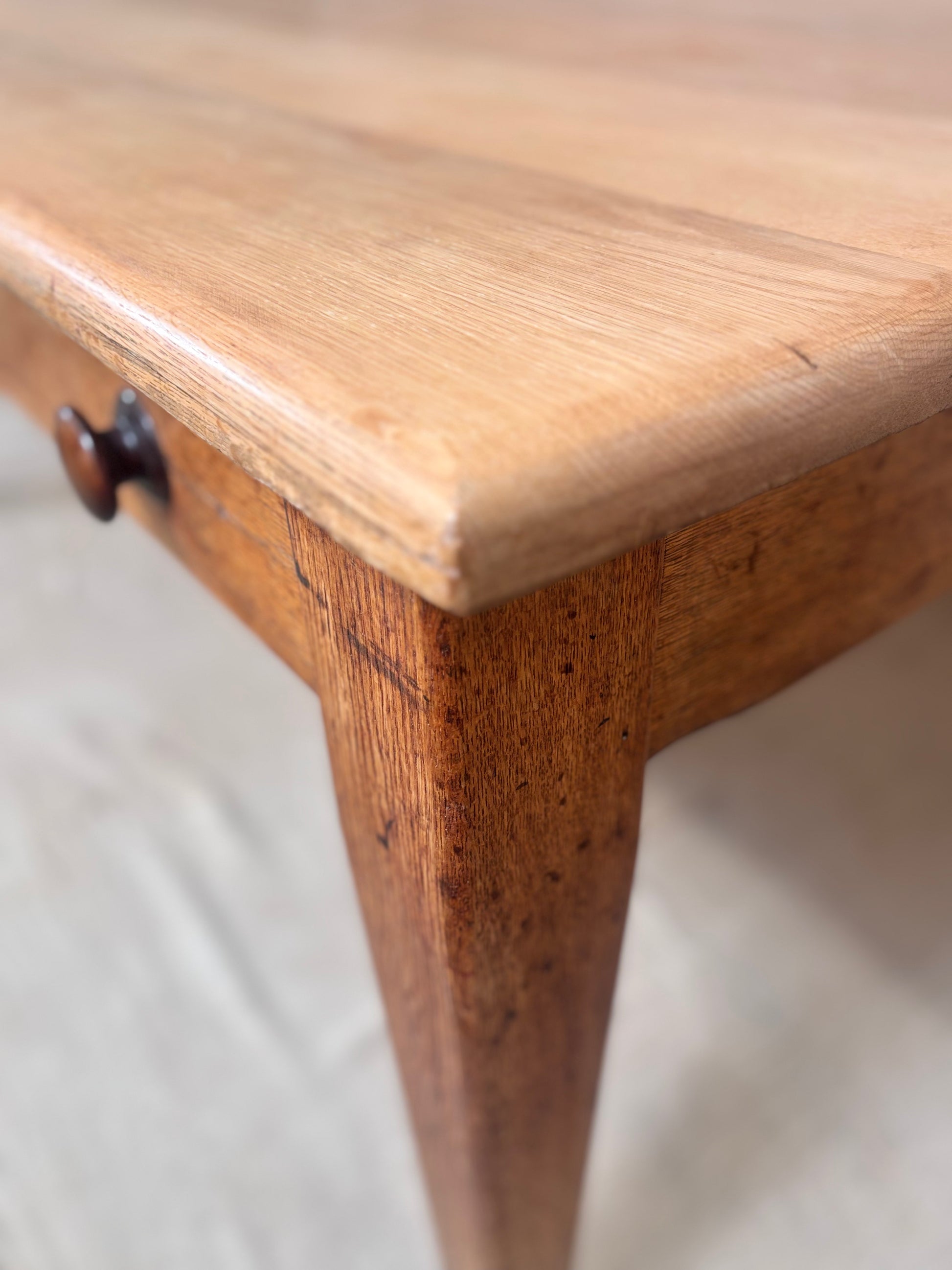Close-up of a wooden table corner showing a light natural wood top, a medium brown leg, and a dark round drawer knob against a light neutral floor.