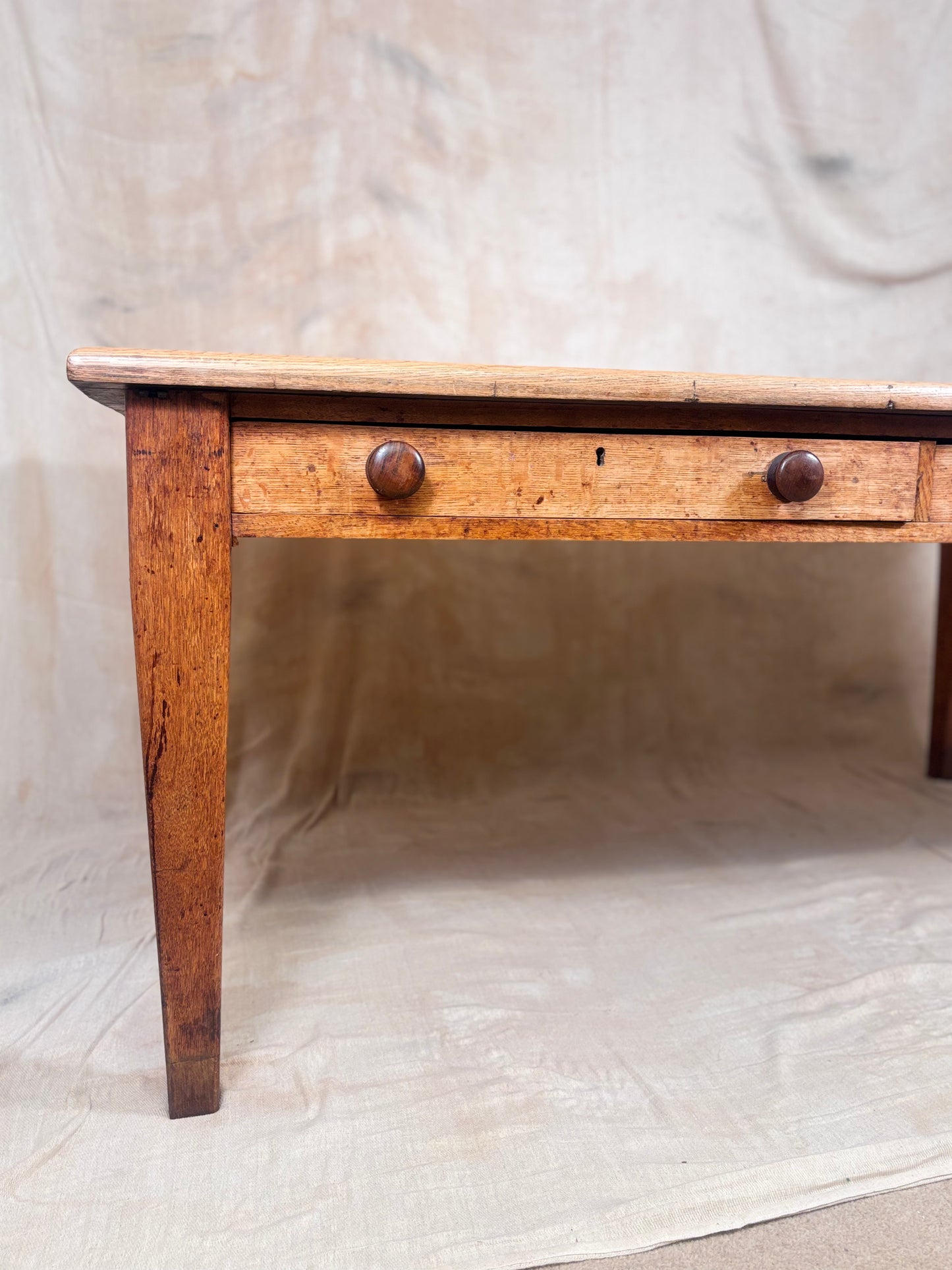 A rustic wooden tablewith a single drawer, two round dark knobs, and a slatted wooden top, set against a draped beige fabric backdrop.