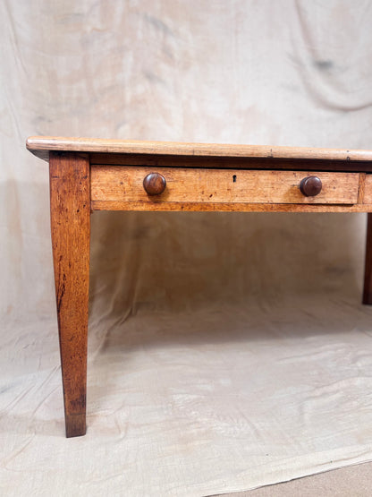 A rustic wooden tablewith a single drawer, two round dark knobs, and a slatted wooden top, set against a draped beige fabric backdrop.