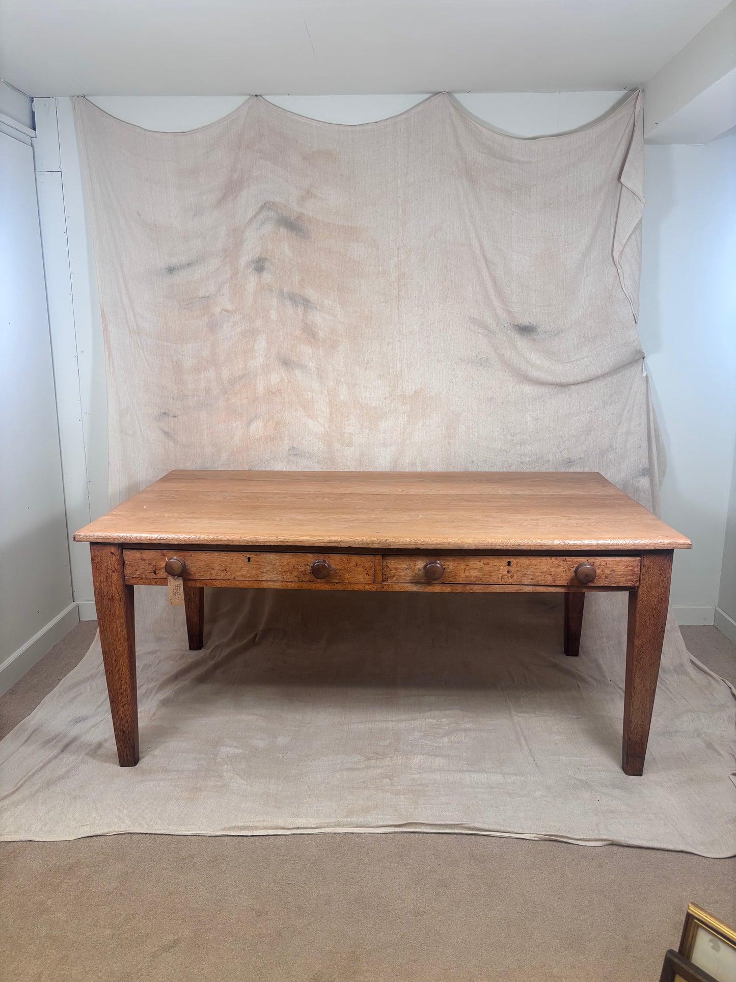 A rustic wooden dining table with a light oak top and medium brown legs, featuring three drawers with dark metal knobs, set against a draped beige fabric backdrop.