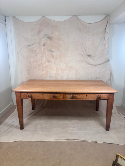 A rustic wooden dining table with a light oak top and medium brown legs, featuring three drawers with dark metal knobs, set against a draped beige fabric backdrop.