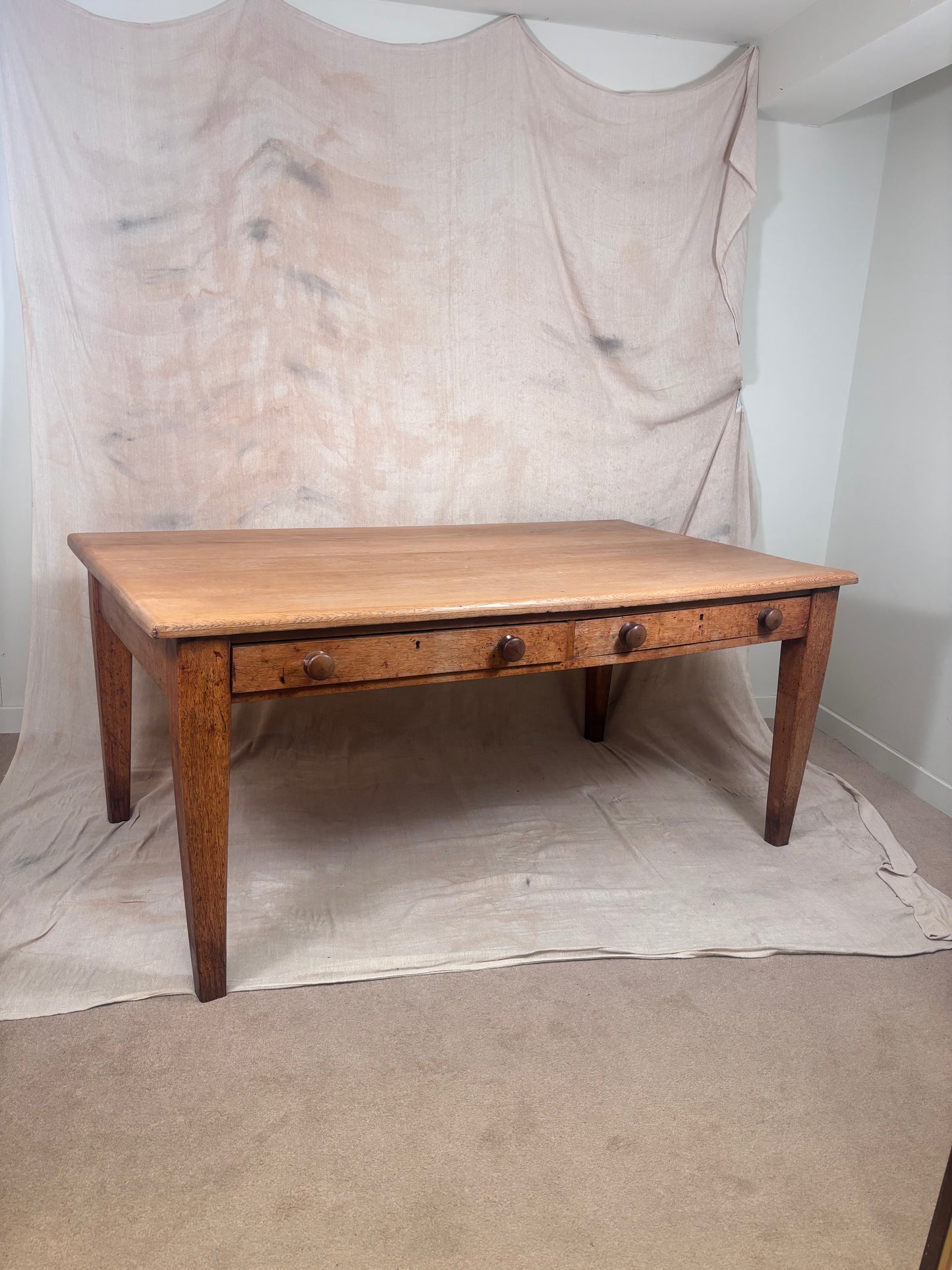 A rustic wooden writing desk with three drawers and round dark knobs, set against a draped beige fabric backdrop.