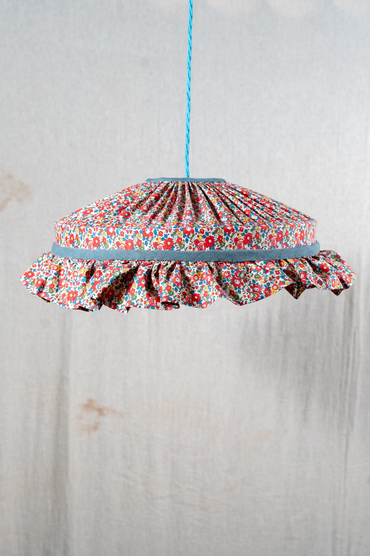 A hanging pendant light shade made of gathered floral fabric with a ruffled edge, suspended by a blue cord against a light textured wall.